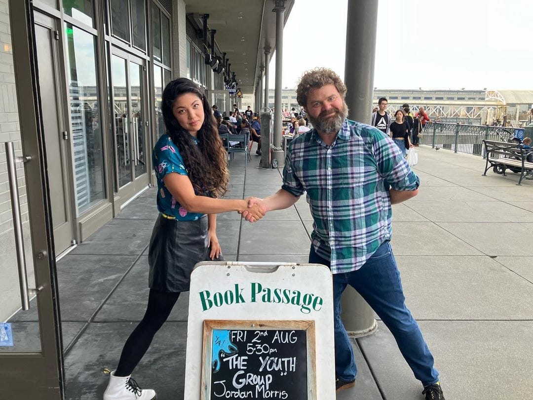 Two writers shaking hands over a "Book Passage" sign.